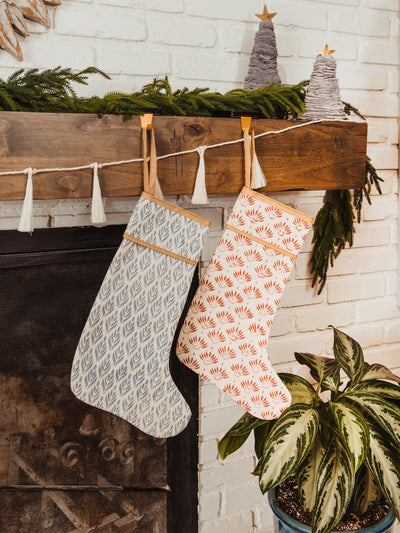 Two handcrafted Christmas stockings, one with a blue leaf pattern and the other with a red leaf pattern, hanging on a rustic wooden mantel adorned with festive greenery, garlands, and decorative mini Christmas trees.
