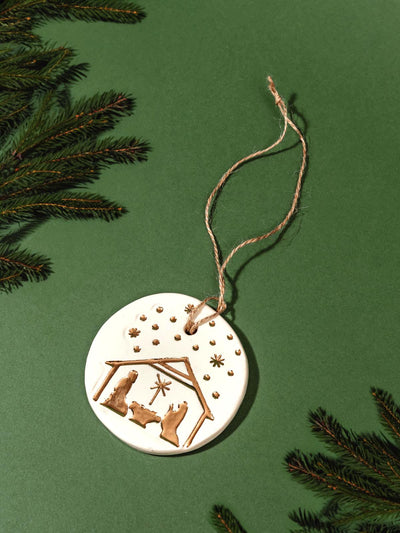 Handmade ceramic nativity ornament with gold accents, featuring a twine hanger, set against a green background with pine branches.