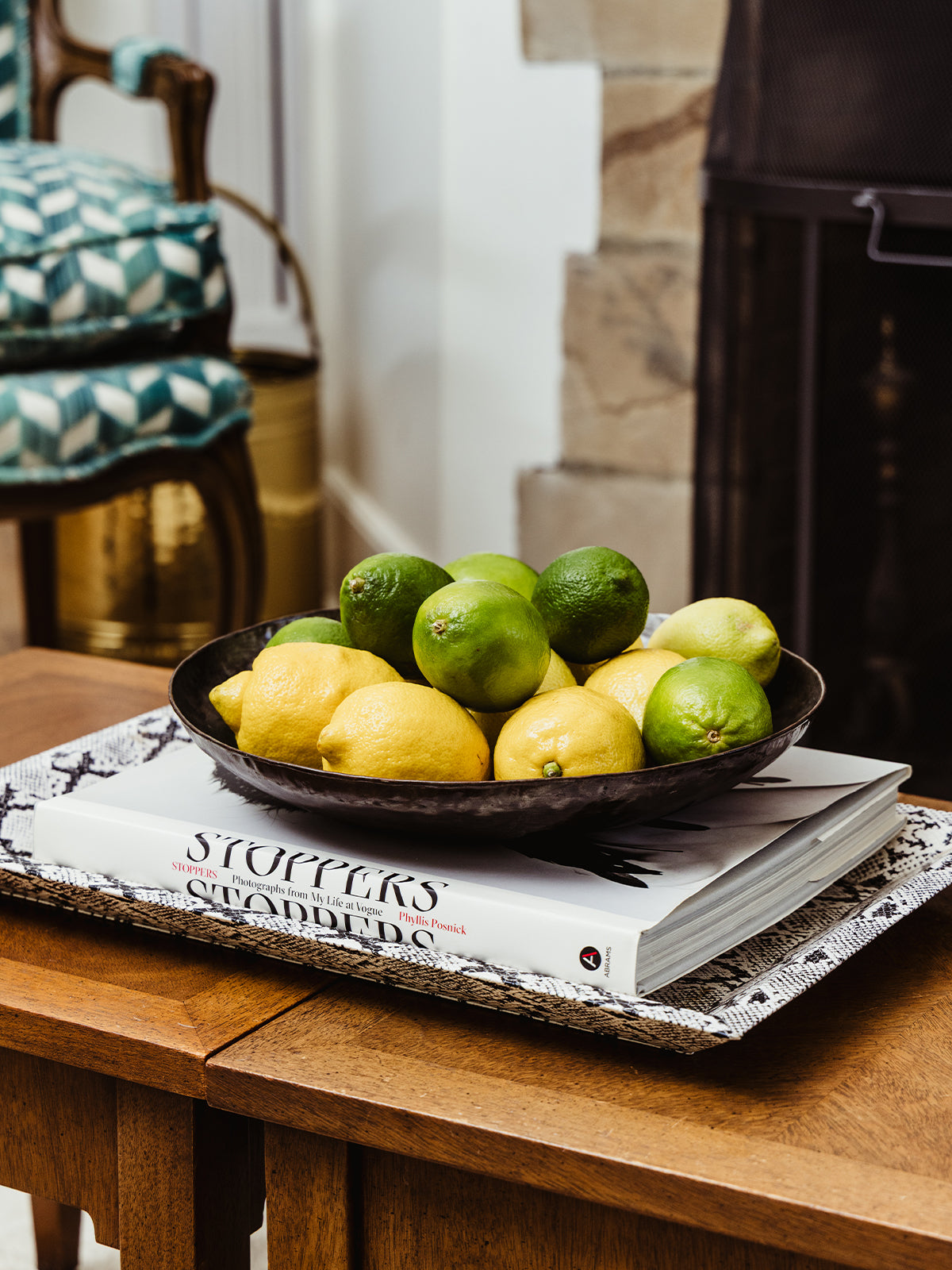 Lemons and limes displayed in a dark bowl atop a fashion book on a wooden side table near a fireplace, styled with cozy, eclectic seating in the background.