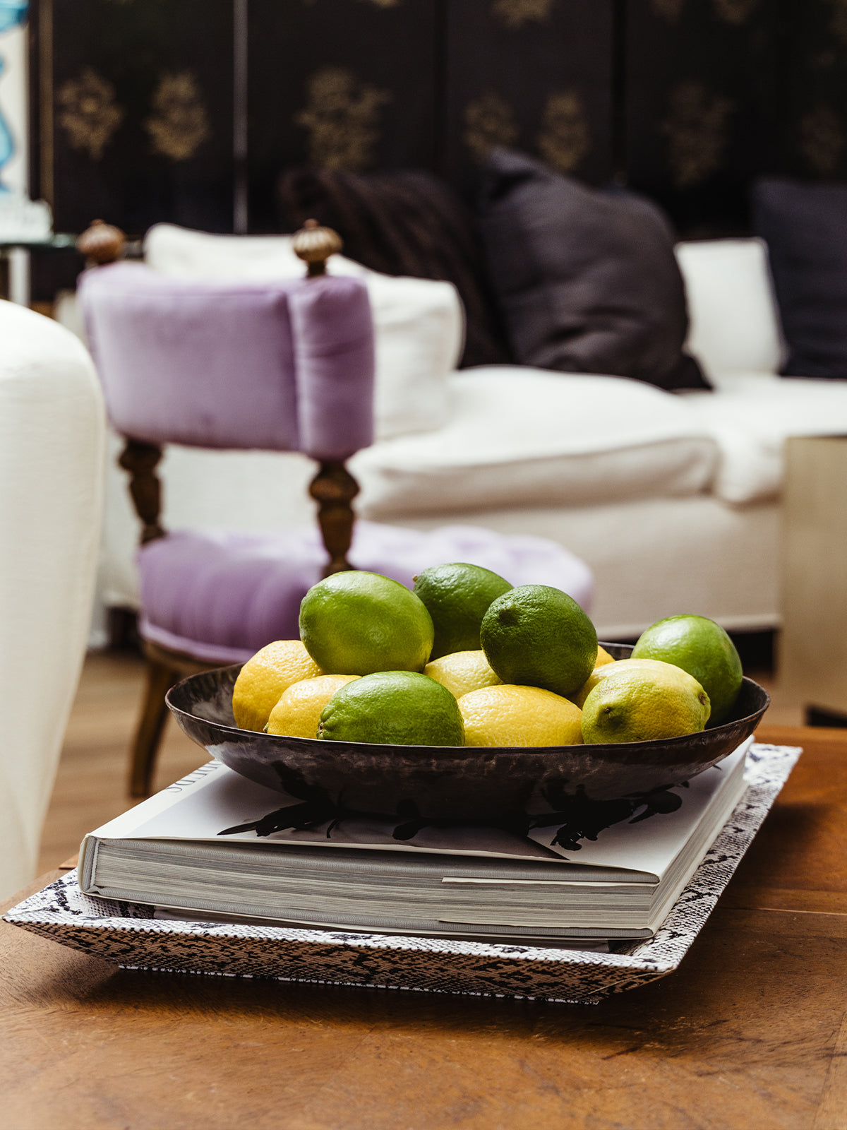 Decorative centerpiece featuring a mix of lemons and limes in a dark metal bowl on top of a white coffee table book, styled in front of a blue and green patterned armchair.