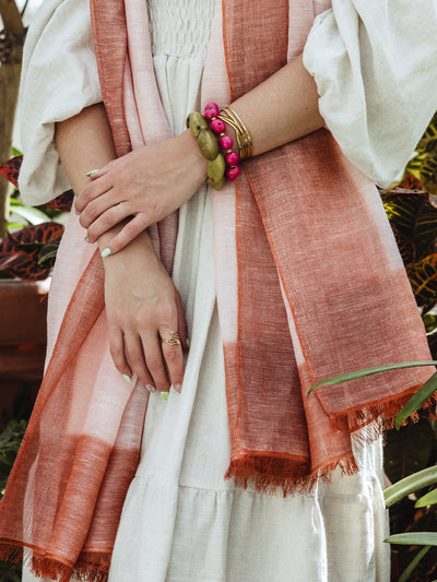 Mid-body shot of a woman in a cream smocked dress and rust-colored scarf, showcasing layered bracelets and rings with a soft natural light background.