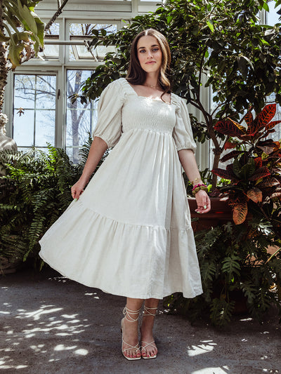 Full-length image of a woman wearing a cream smocked midi dress with puff sleeves and strappy heels, posing in a lush greenhouse.