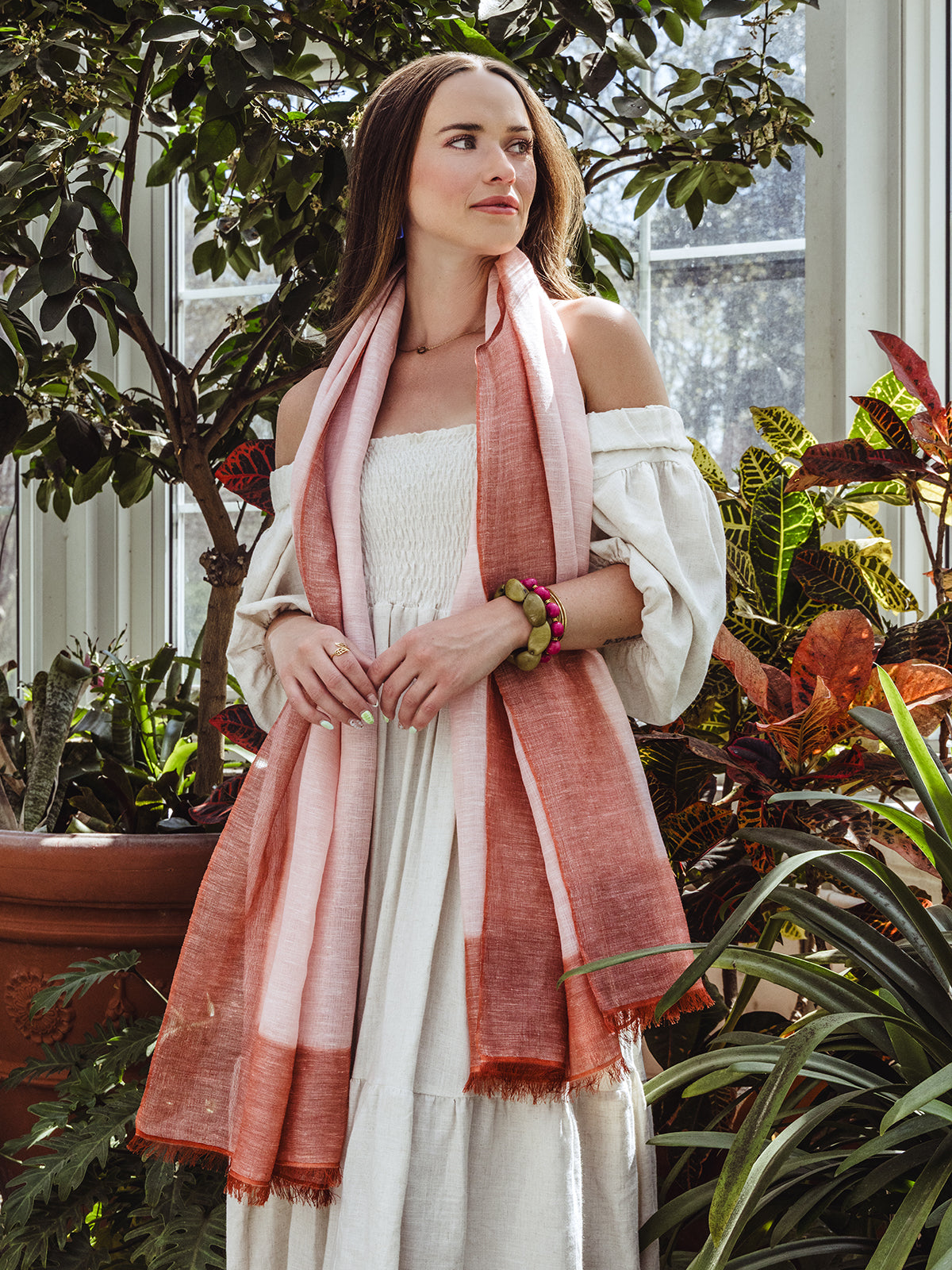 Woman in an off-shoulder cream dress and a peach and rust gradient scarf, standing amidst indoor greenery, accessorized with chunky bracelets and a simple necklace.