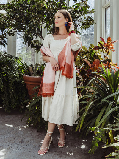 Full-body photo of a woman wearing a cream dress, rust scarf, and bold earrings, standing confidently in a greenhouse surrounded by tropical plants.