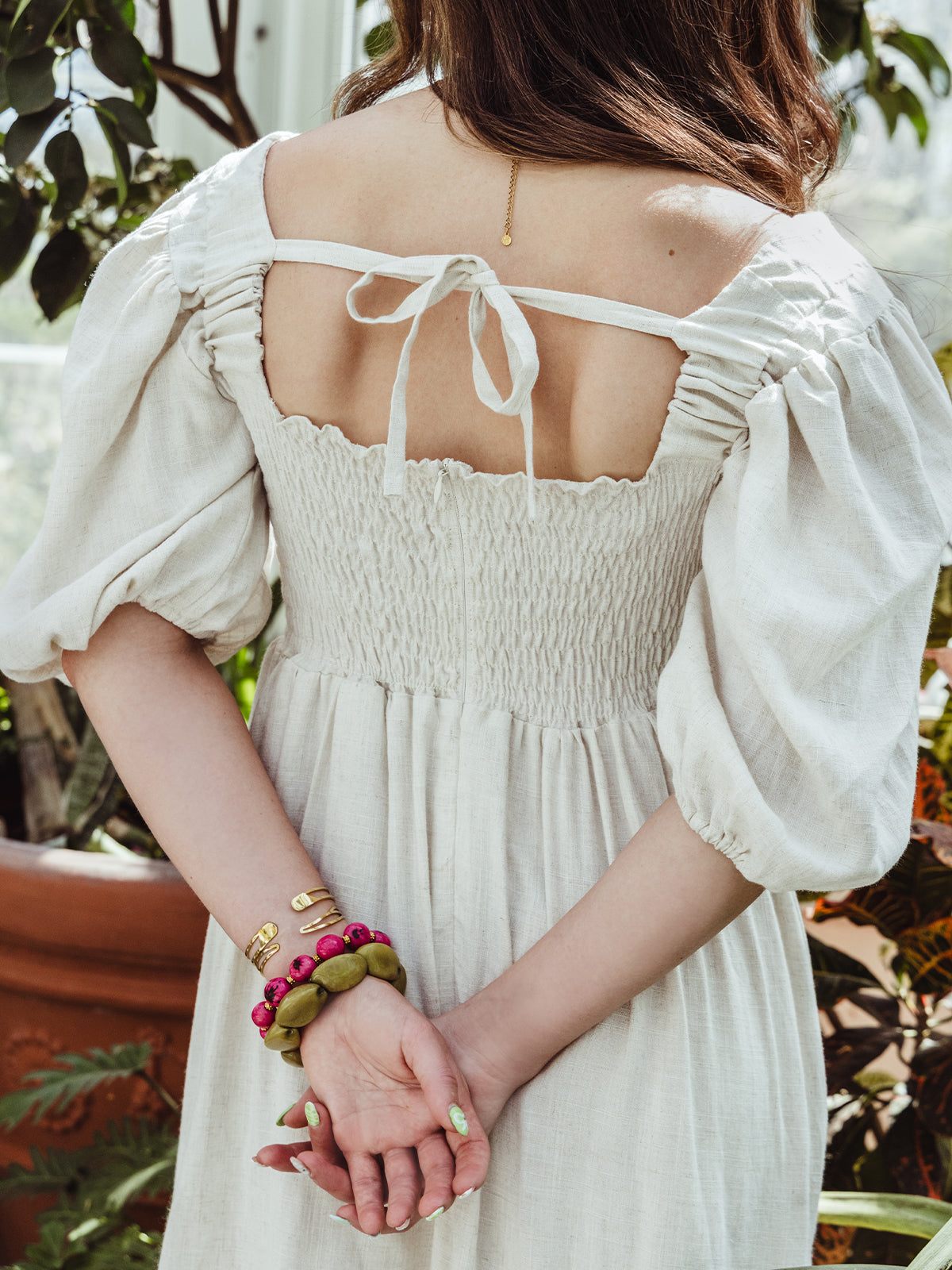 Back view of a woman in a smocked cream dress with a tied bow detail and layered bracelets, holding her hands gently behind her back.