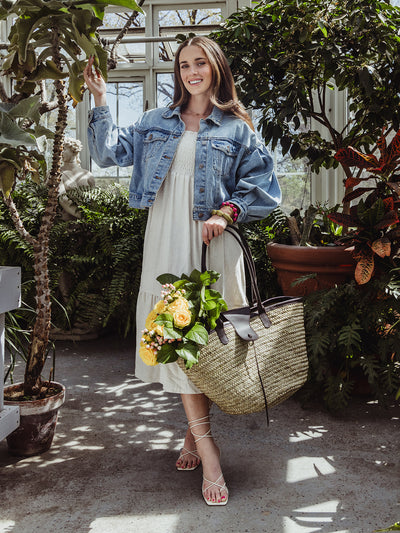 Woman wearing a cream midi dress with a denim jacket and straw tote bag filled with yellow roses, standing joyfully in a sunlit greenhouse.
