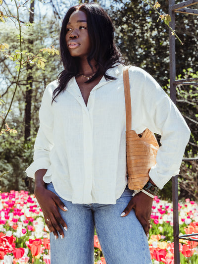 Woman wearing a loose white button-up blouse and light wash jeans, standing in front of vibrant spring tulips with a natural woven shoulder bag.