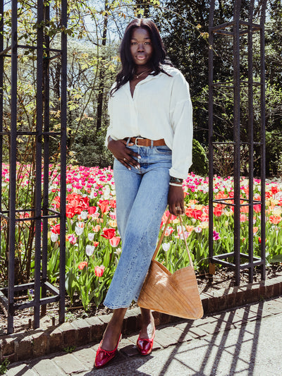 Full-body shot of a woman in a white blouse and jeans with red shoes, standing confidently in a flower garden holding a woven tan handbag.