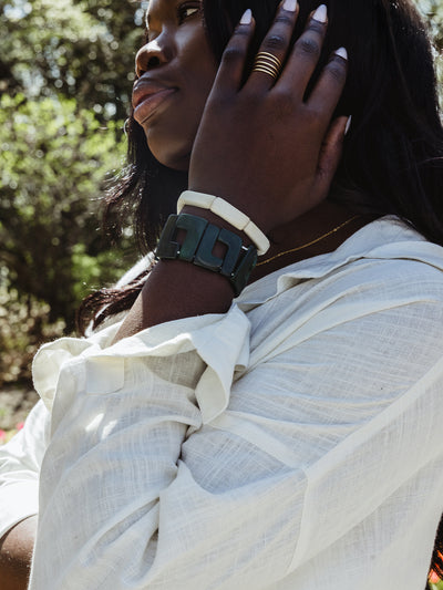 Close-up of a woman’s wrist and hand, showcasing stacked bracelets in black and ivory tones with a gold ring, set against a softly lit garden background.