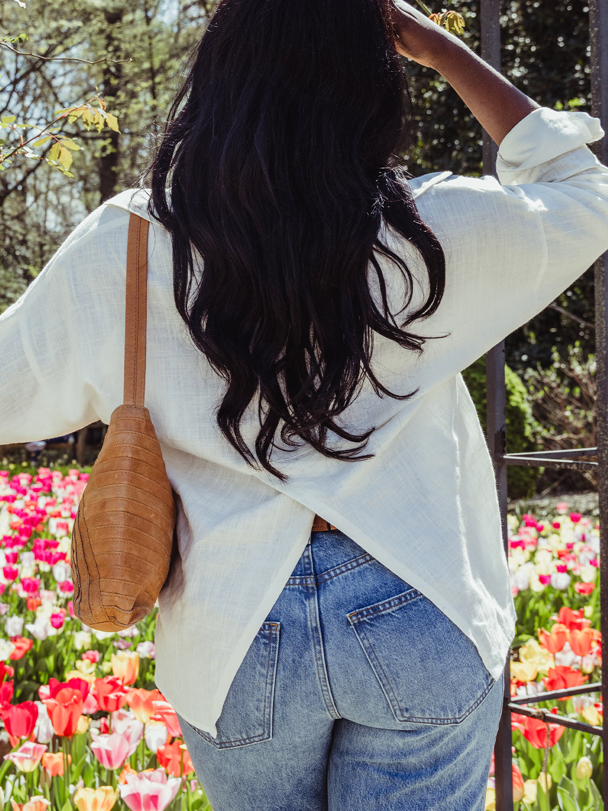 Back view of a woman with long dark hair wearing a white blouse with a crisscross hem and high-waisted jeans, standing before blooming tulips.