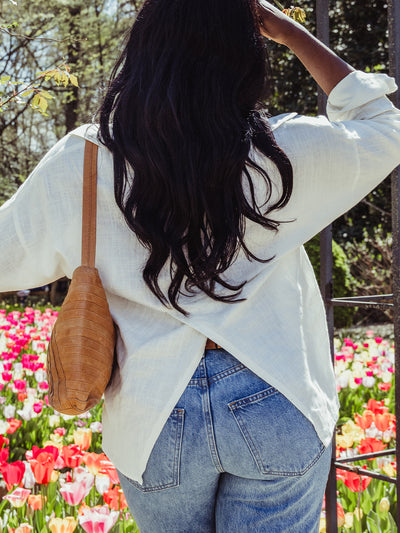 Back view of a woman with long dark hair wearing a white blouse with a crisscross hem and high-waisted jeans, standing before blooming tulips.