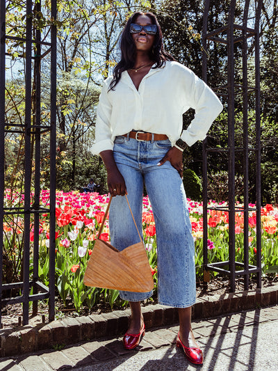 Woman standing confidently in front of colorful tulips, wearing red flats, light-wash denim, and a white blouse tucked in with a brown belt.