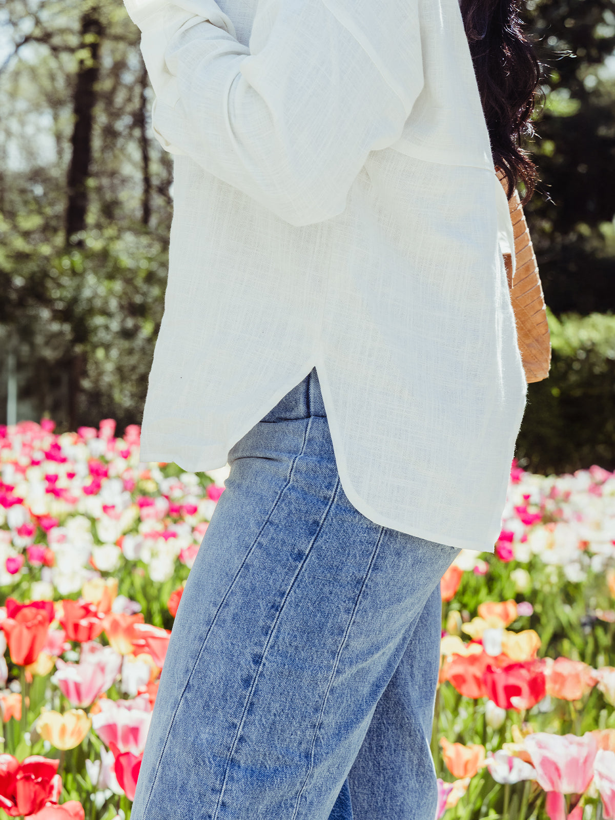 Side view of a woman in a white blouse with a side slit hem, paired with high-rise jeans, standing beside blooming tulips in a spring garden.