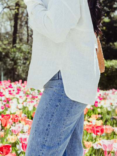 Side view of a woman in a white blouse with a side slit hem, paired with high-rise jeans, standing beside blooming tulips in a spring garden.