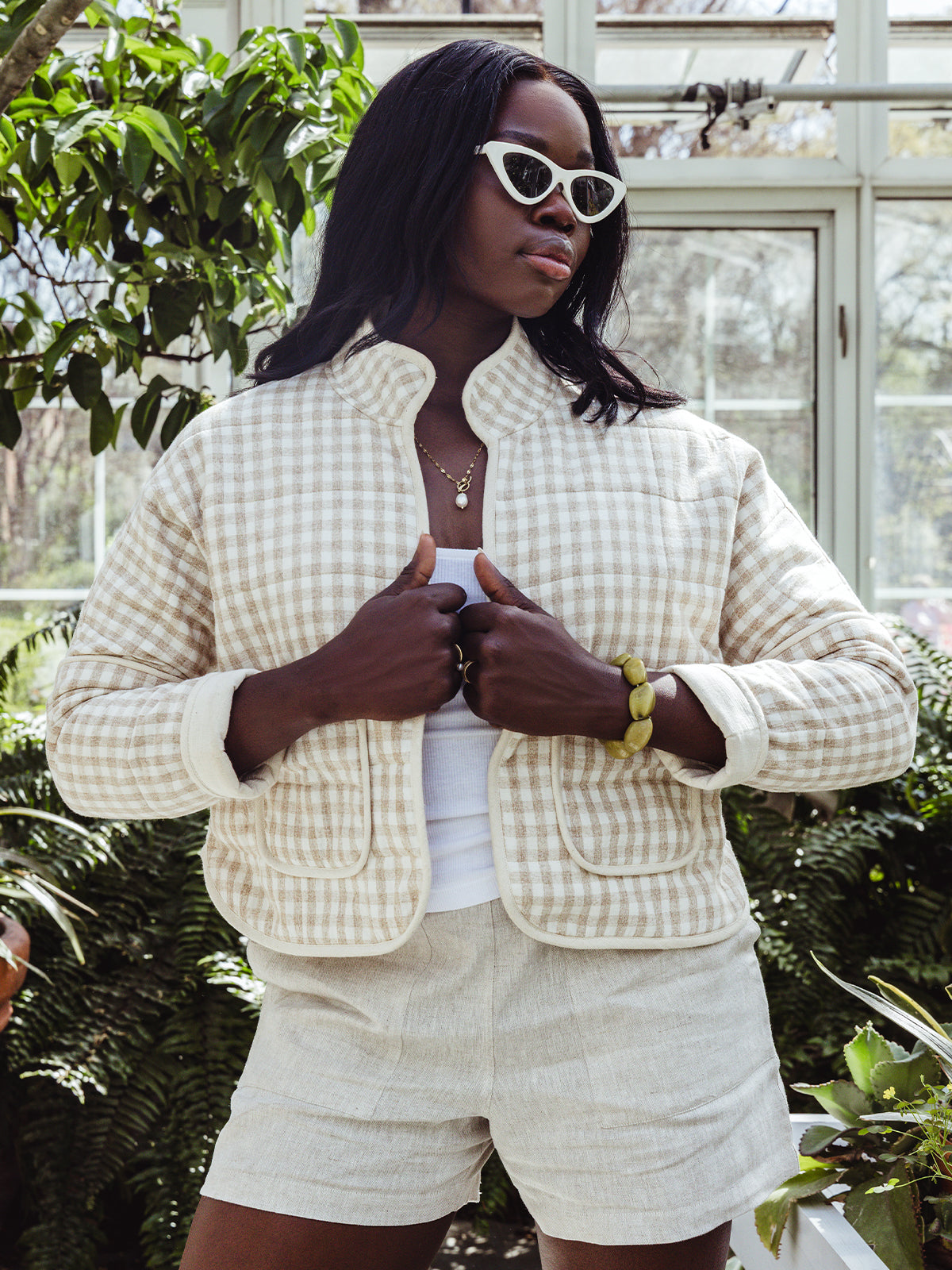 A woman wearing a cream and beige gingham quilted jacket over a white tank top and linen shorts stands in a sunlit greenhouse. She styles the look with white cat-eye sunglasses, a gold necklace, and a chunky green bracelet.