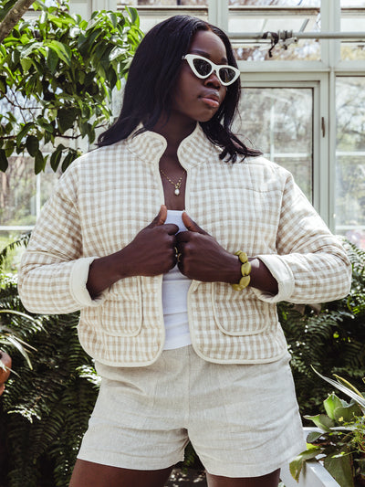 A woman wearing a cream and beige gingham quilted jacket over a white tank top and linen shorts stands in a sunlit greenhouse. She styles the look with white cat-eye sunglasses, a gold necklace, and a chunky green bracelet.