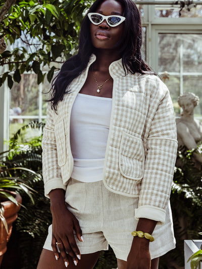 Close-up of a woman in a quilted gingham jacket and white ribbed tank top. She wears cat-eye sunglasses, a delicate gold necklace with a pearl charm, and rests her hand casually in the pocket of her high-waisted linen shorts.