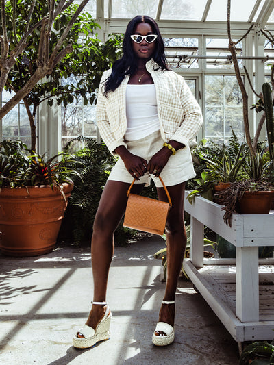 Full-body view of a woman in a light, summery outfit featuring a gingham quilted jacket, white tank top, linen shorts, and espadrille wedges. She accessorizes with a tan woven handbag, cat-eye sunglasses, and bold green bracelet in a greenhouse setting.