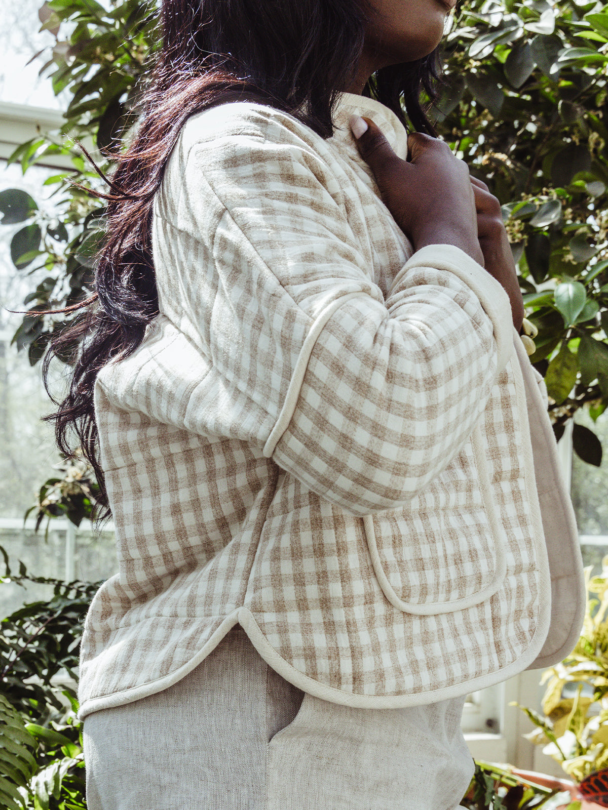 Side view of a woman wearing a beige gingham quilted jacket with curved hem detailing. Her hand gently holds the jacket closed as sunlight highlights the jacket's texture and stitching.