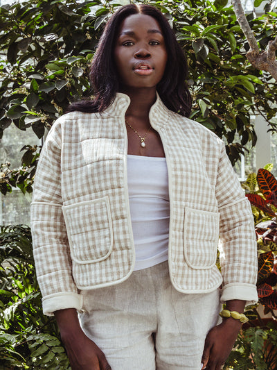 Medium close-up of a woman standing in front of lush greenery, wearing a cream and tan gingham jacket and light linen shorts. She pairs the look with a gold pendant necklace and minimal makeup.