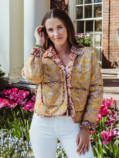 Woman standing in front of a flower garden wearing a colorful floral quilted jacket with red and yellow tones, white jeans, and a tank top.