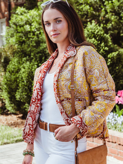 Woman standing outdoors in a patterned floral jacket with red trim, white top and jeans, accessorized with a brown leather crossbody bag and white sunglasses.