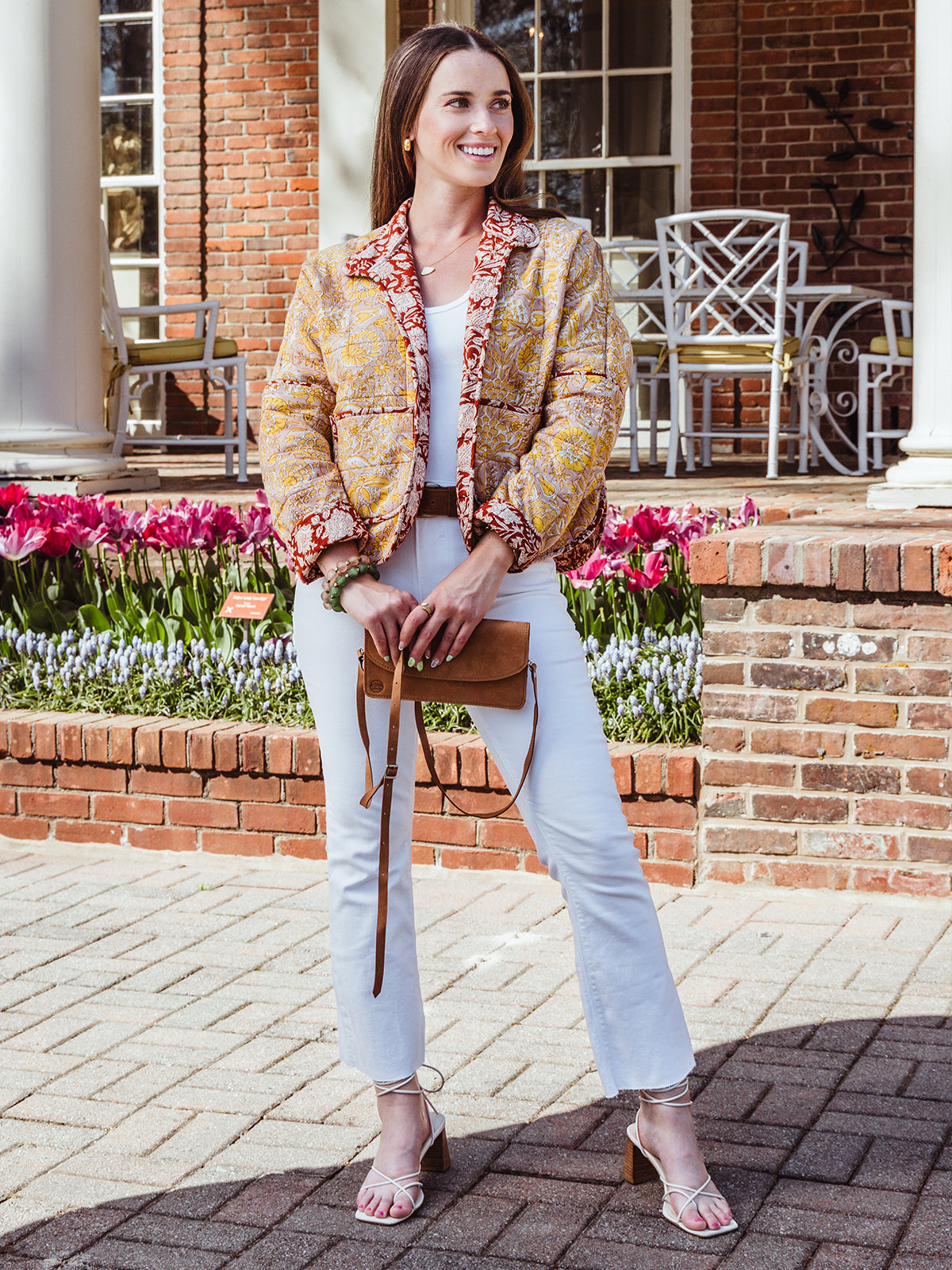 Full-body view of a woman wearing a yellow and red floral quilted jacket, white jeans, strappy heels, and a brown leather wristlet purse, posing in front of tulips and a brick building.