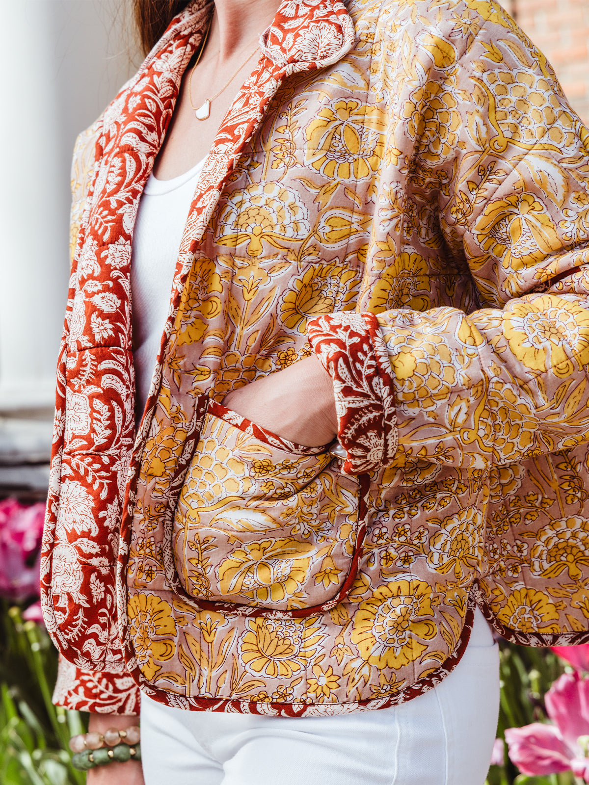 Close-up of a woman’s hand in the pocket of a floral block print quilted jacket, showing the red trim, detailed yellow floral pattern, and cozy stitching.