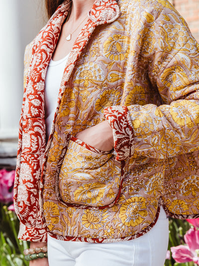 Close-up of a woman’s hand in the pocket of a floral block print quilted jacket, showing the red trim, detailed yellow floral pattern, and cozy stitching.