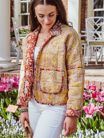 Woman wearing a floral quilted jacket in pink, yellow, and red tones, looking down with a soft smile while standing in front of blooming tulips.