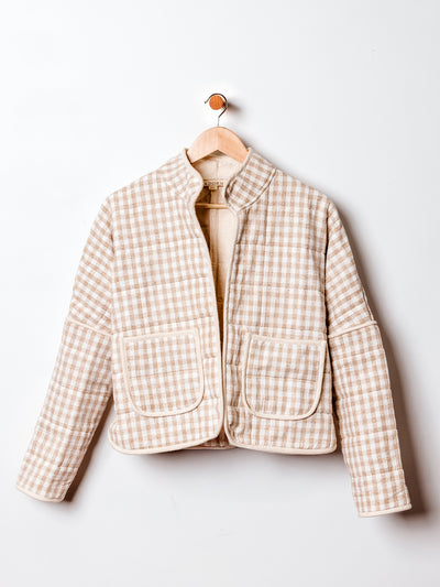A beige and white gingham quilted jacket hangs on a wooden hanger against a white wall. The jacket features a mandarin collar, two large front patch pockets, and a soft, padded texture with a structured silhouette. The neutral tones give it a cozy, classic appeal.