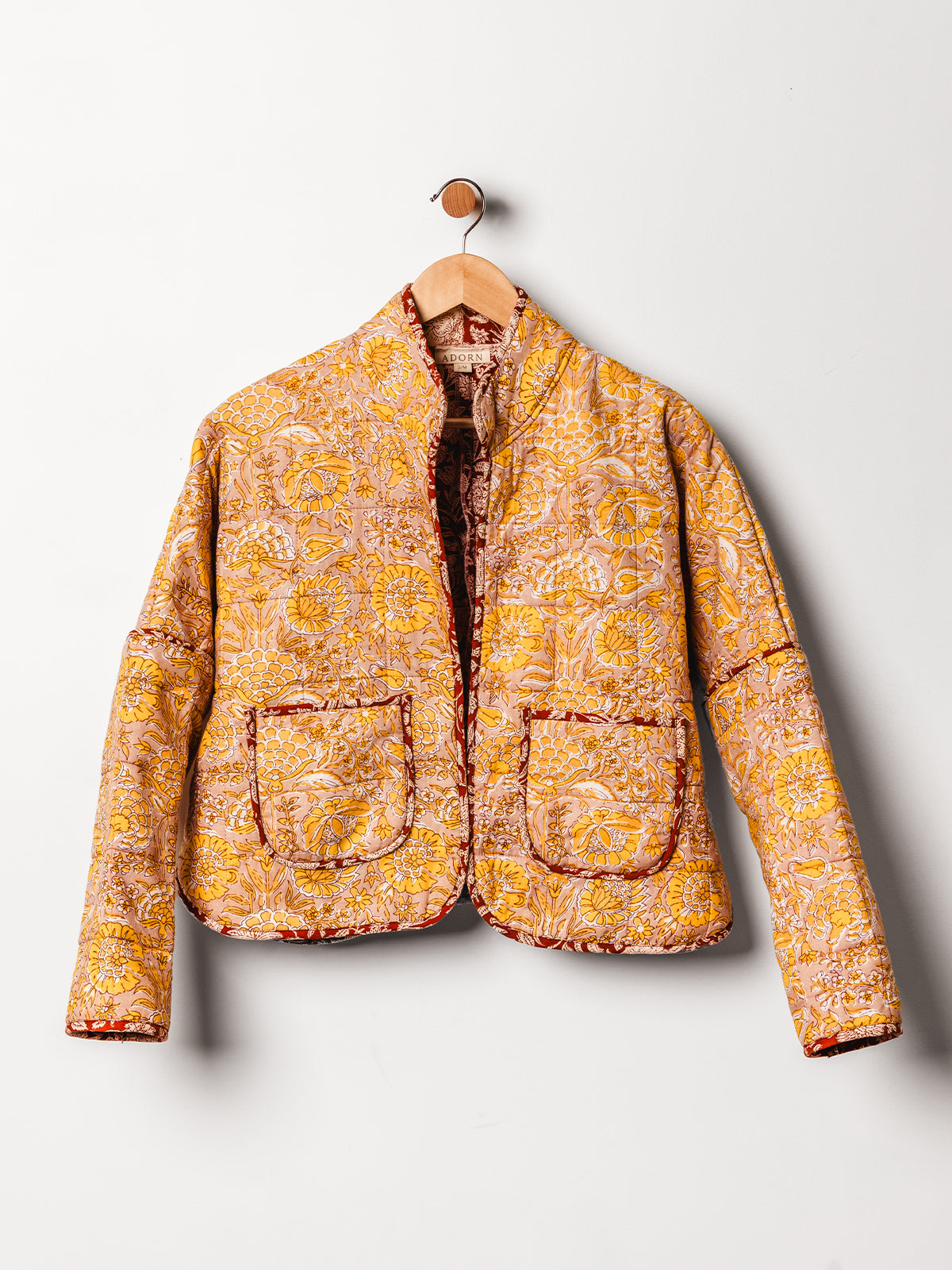 A vibrant yellow and orange floral quilted jacket is displayed on a wooden hanger against a white wall. The jacket has a stand-up collar, intricate hand-stitched detailing, and two large patch pockets trimmed in red. Its warm colors and detailed pattern evoke a handcrafted, artisanal style.