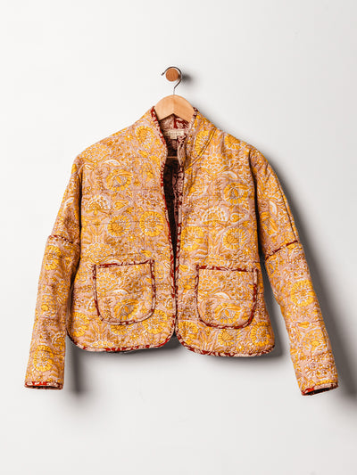 A vibrant yellow and orange floral quilted jacket is displayed on a wooden hanger against a white wall. The jacket has a stand-up collar, intricate hand-stitched detailing, and two large patch pockets trimmed in red. Its warm colors and detailed pattern evoke a handcrafted, artisanal style.