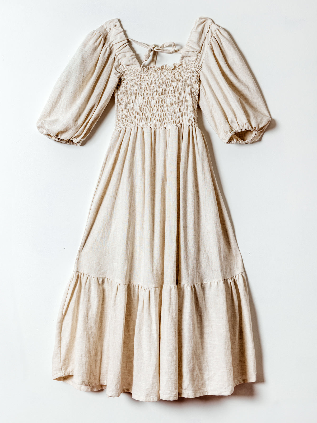 A soft beige midi dress hangs flat against a white background. The dress features puffed half sleeves, a smocked bodice with a subtle ruffled neckline, and a flowy tiered skirt. A tie at the back neckline adds a delicate detail to the feminine and airy silhouette.
