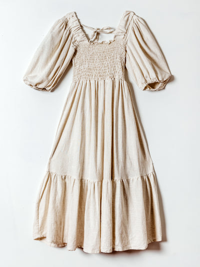 A soft beige midi dress hangs flat against a white background. The dress features puffed half sleeves, a smocked bodice with a subtle ruffled neckline, and a flowy tiered skirt. A tie at the back neckline adds a delicate detail to the feminine and airy silhouette.