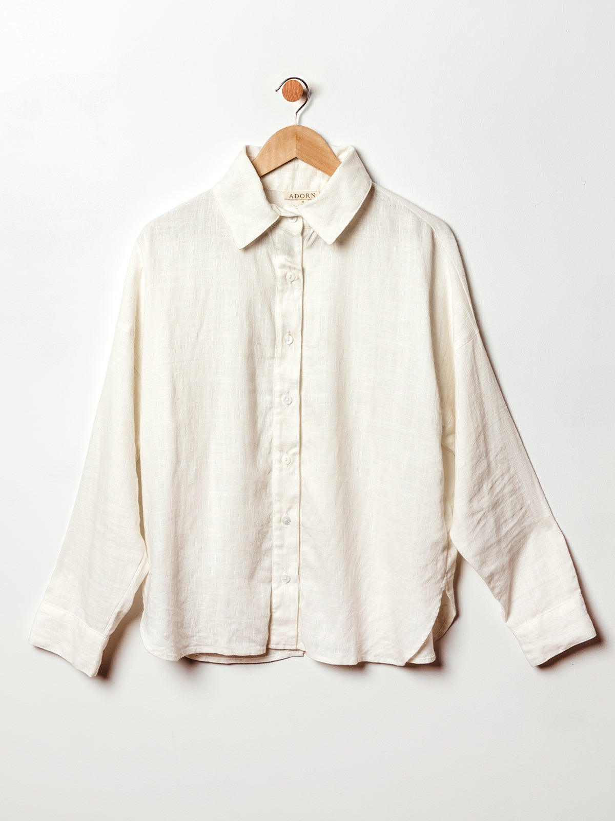 A long-sleeve button-up shirt in a soft off-white linen fabric hangs on a wooden hanger against a white wall. The shirt features a classic collar, relaxed fit, and subtle texture, with a gently curved hem and cuffed sleeves for a timeless, breathable look.