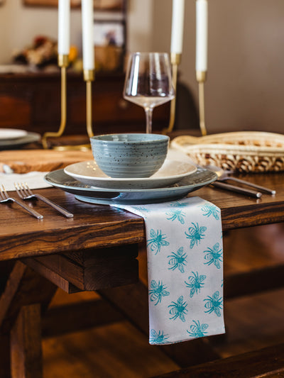 Hand-printed white cotton napkin with teal bee pattern draped over a rustic wood dining table, styled with stoneware dinnerware, wine glass, and brass candle holders — fair trade table décor.