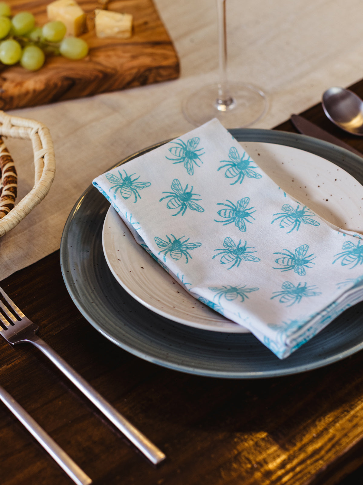 Folded white cotton napkin with teal bee block print displayed on speckled ceramic dinner plate at an elegant dining setup with charcuterie board and woven basket — artisan-made and sustainable.