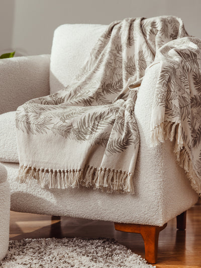 Neutral boho throw blanket with fern leaf design and fringe draped over a white boucle armchair, cozy living room decor inspiration.