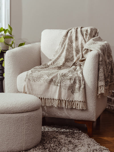 Handwoven botanical print throw blanket with tassel fringe styled on a cream armchair and ottoman, sustainable home decor accent.