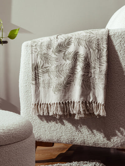 Eco-friendly beige throw blanket with leafy botanical pattern and fringed edges displayed on modern boucle chair, ethical living room styling.