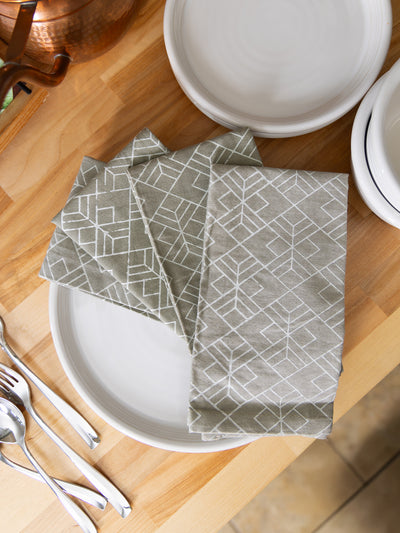 Set of handmade cloth napkins in a neutral gray geometric pattern, neatly folded on a white plate with silver flatware on a wooden kitchen counter — sustainable artisan-made table linens.