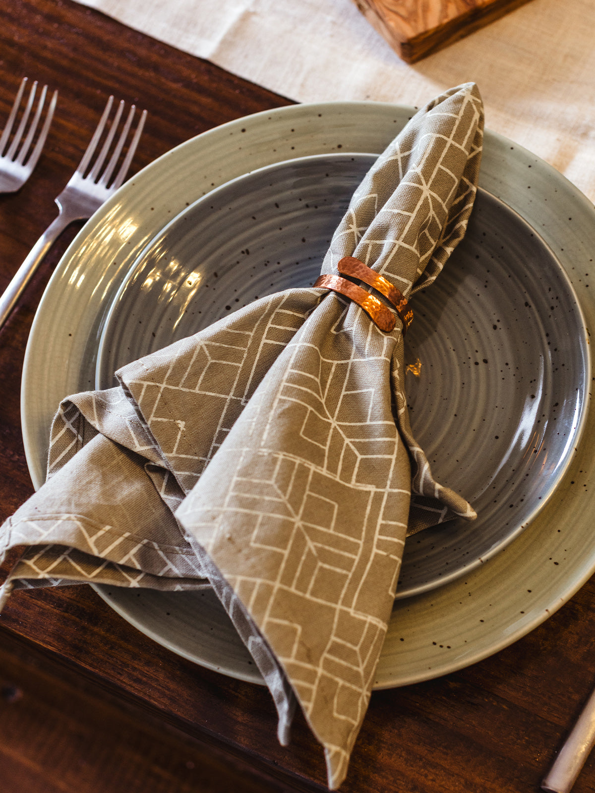 Handcrafted neutral cotton napkin with modern geometric print, styled with a hammered copper napkin ring on a speckled ceramic dinner plate — ethical, fair trade table décor.
