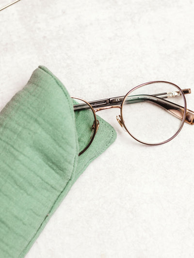 Handmade mint green muslin eyeglass case with gold-rimmed glasses partially inside, on a light marble background.