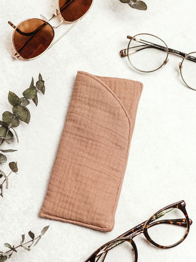 Soft blush color handmade muslin eyeglass case surrounded by various eyeglasses and eucalyptus leaves on a white marble surface.