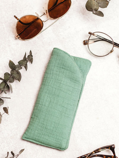 Handmade muslin eyeglass case in mint green with eyeglasses nearby, surrounded by sunglasses and eucalyptus leaves on a white background.