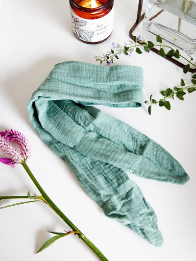 Mint green fabric wrap headband displayed on a white surface alongside a lit candle, flowers, and greenery, highlighting its soft texture.