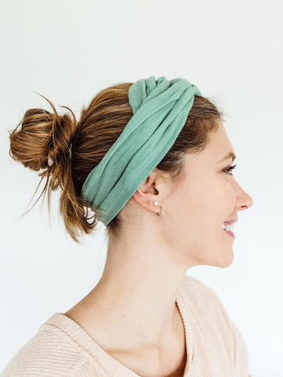Side profile of a woman wearing a mint green fabric wrap headband, styled with a casual messy bun, smiling and looking to the side.