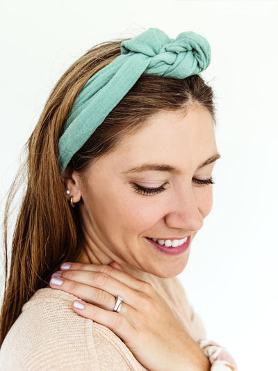 Woman smiling and looking down while wearing a mint green fabric wrap headband tied in a knot at the top, paired with a casual sweater.