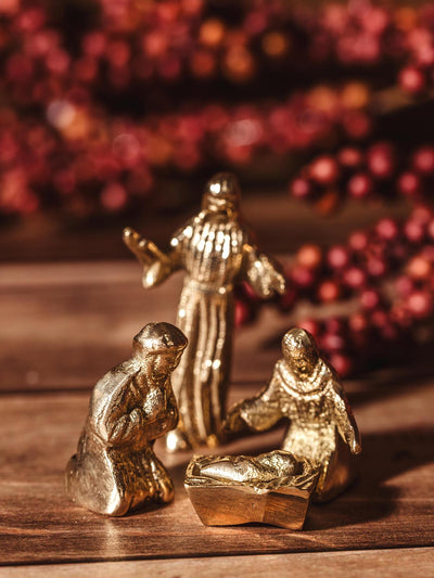 Handcrafted brass nativity figures of baby Jesus in a manger, Mary, Joseph, and a shepherd, set against a festive background.